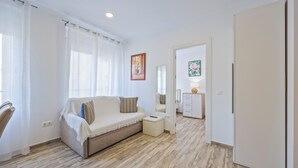 A modern living room with a beige sofa, a small ottoman, and a white cabinet. There is a door leading to another room and a painting on the wall. - Apartamentos Chorrol (Benidorm)