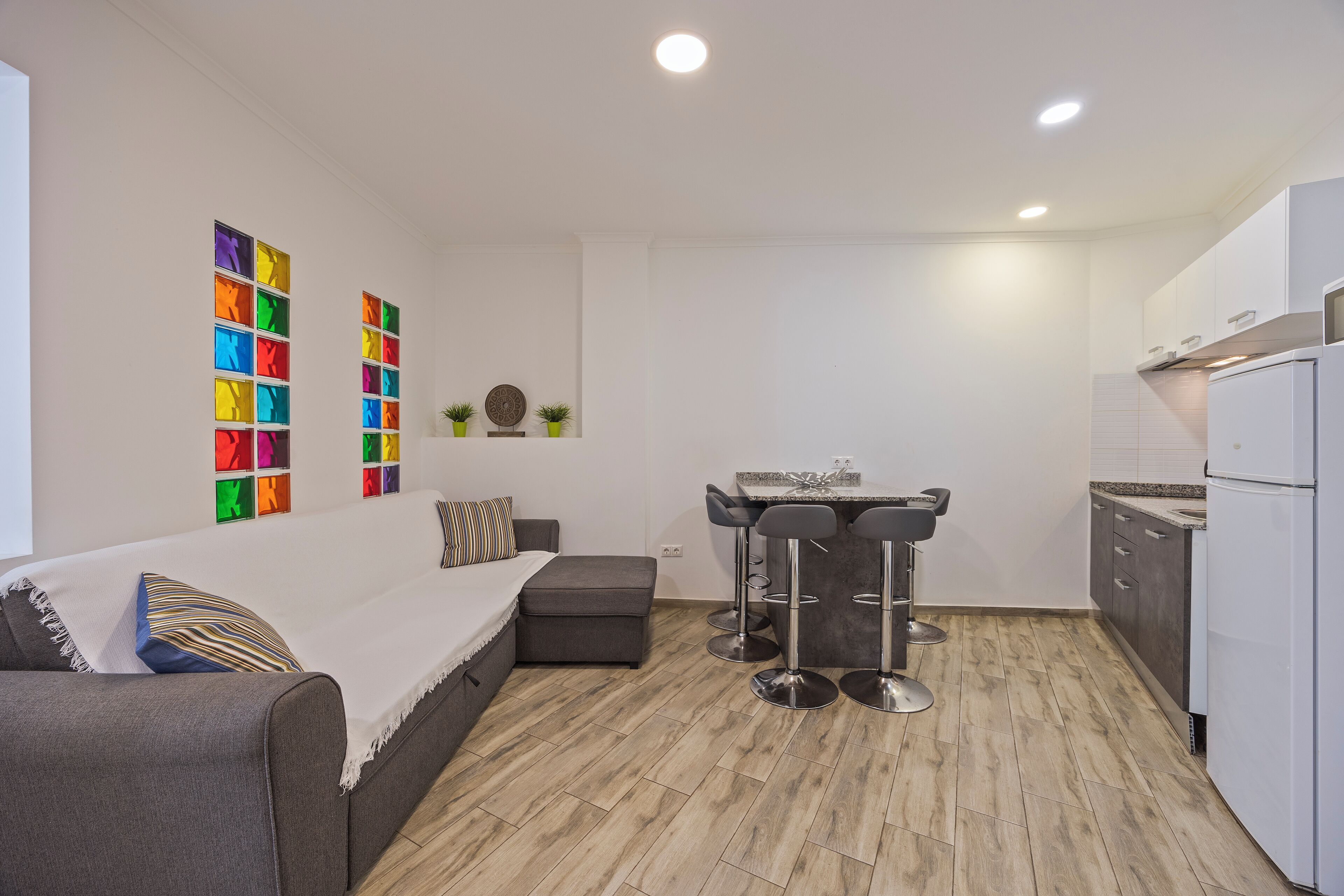 A compact living space with a sofa, a dining table with bar stools, a kitchenette, and a wall with colorful artwork.