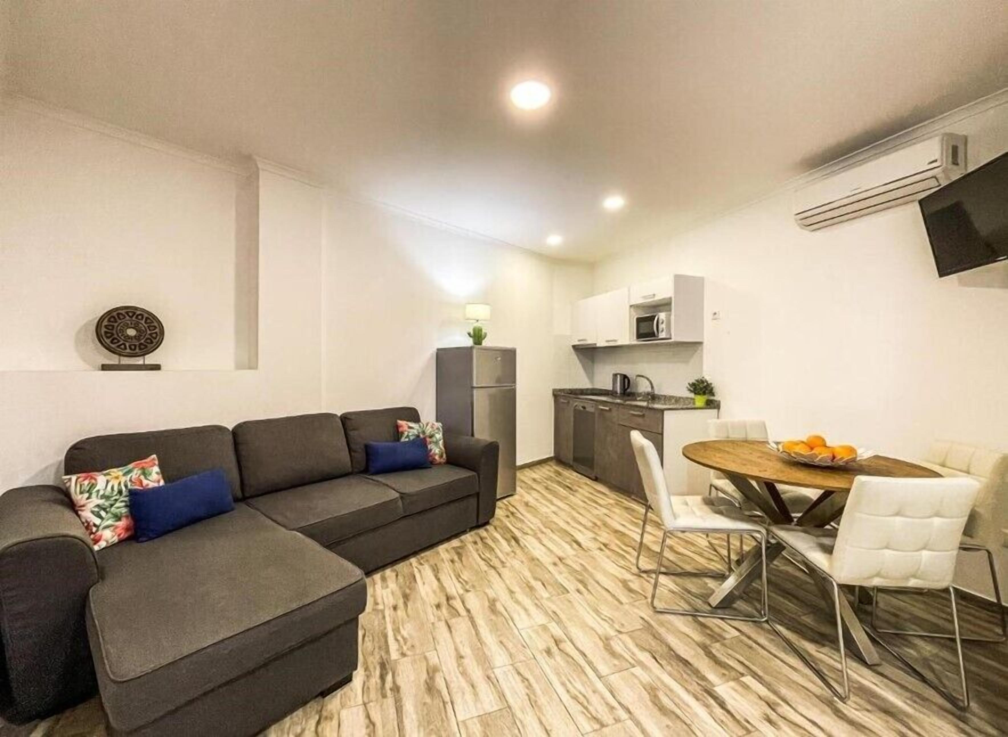 Una sala moderna con un sofá seccional gris, una mesa de comedor con sillas blancas y una cocina con armarios blancos y un microondas.