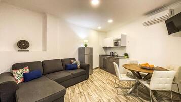 Una sala moderna con un sofá seccional gris, una mesa de comedor con sillas blancas y una cocina con armarios blancos y un microondas.