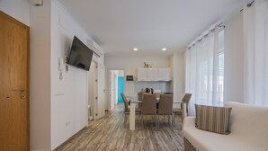 A modern living room with a sofa, a dining area with a table and chairs, a kitchen with cabinets and a microwave, and a TV mounted on the wall. - Apartamentos Chorrol (Benidorm)