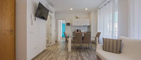 A modern living room with a sofa, a dining area with a table and chairs, a kitchen with cabinets and a microwave, and a TV mounted on the wall.
