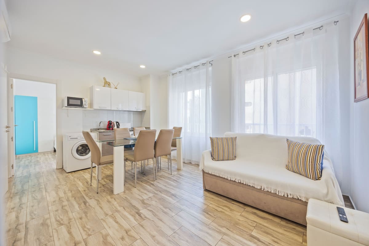 A modern living room with a sofa, a dining area with chairs, a washing machine, and a microwave.