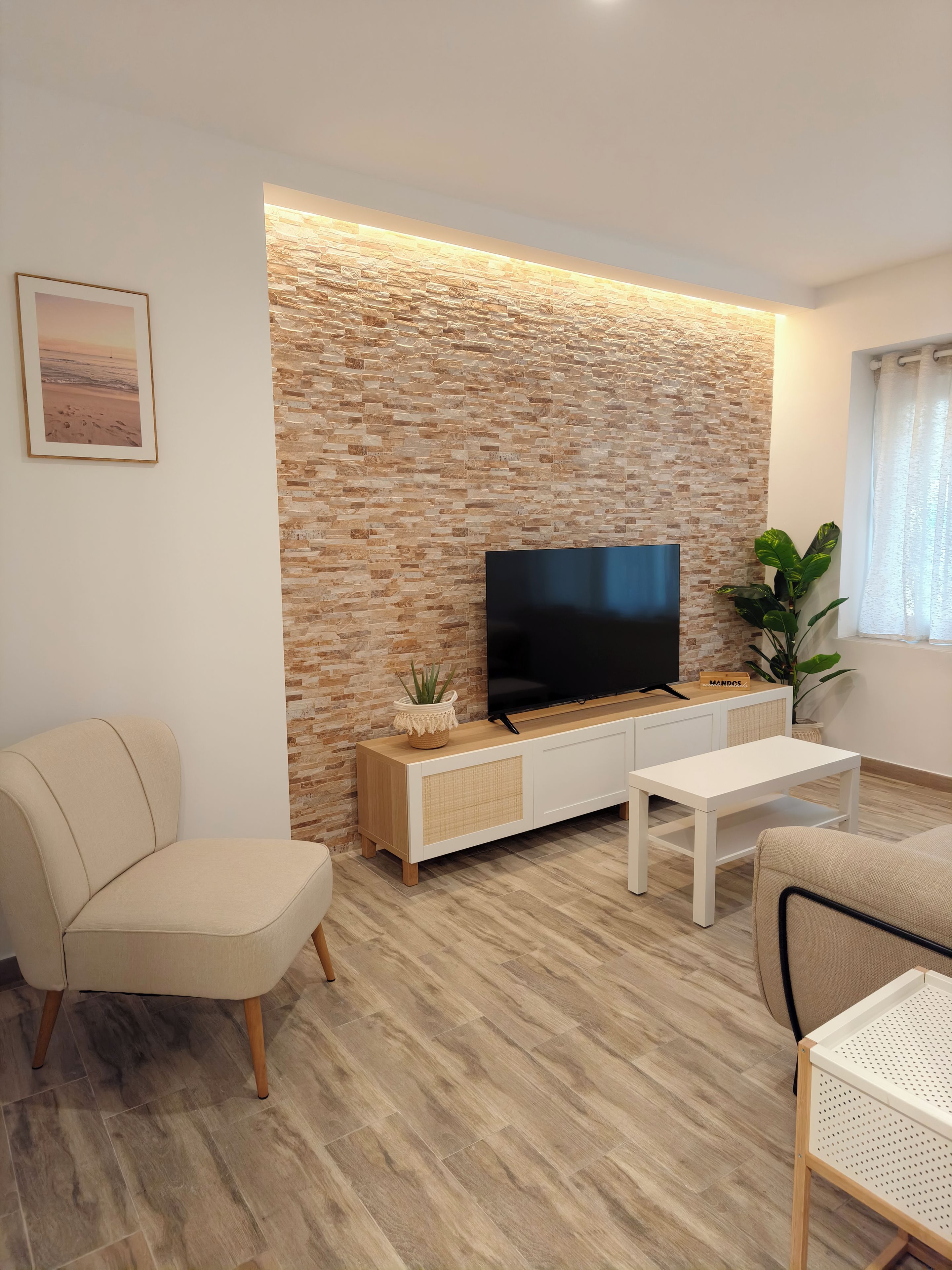 Ein modernes Wohnzimmer mit einer Wand aus Stein, einem Flachbildfernseher, einer weißen Bank und einem beigefarbenen Sessel.