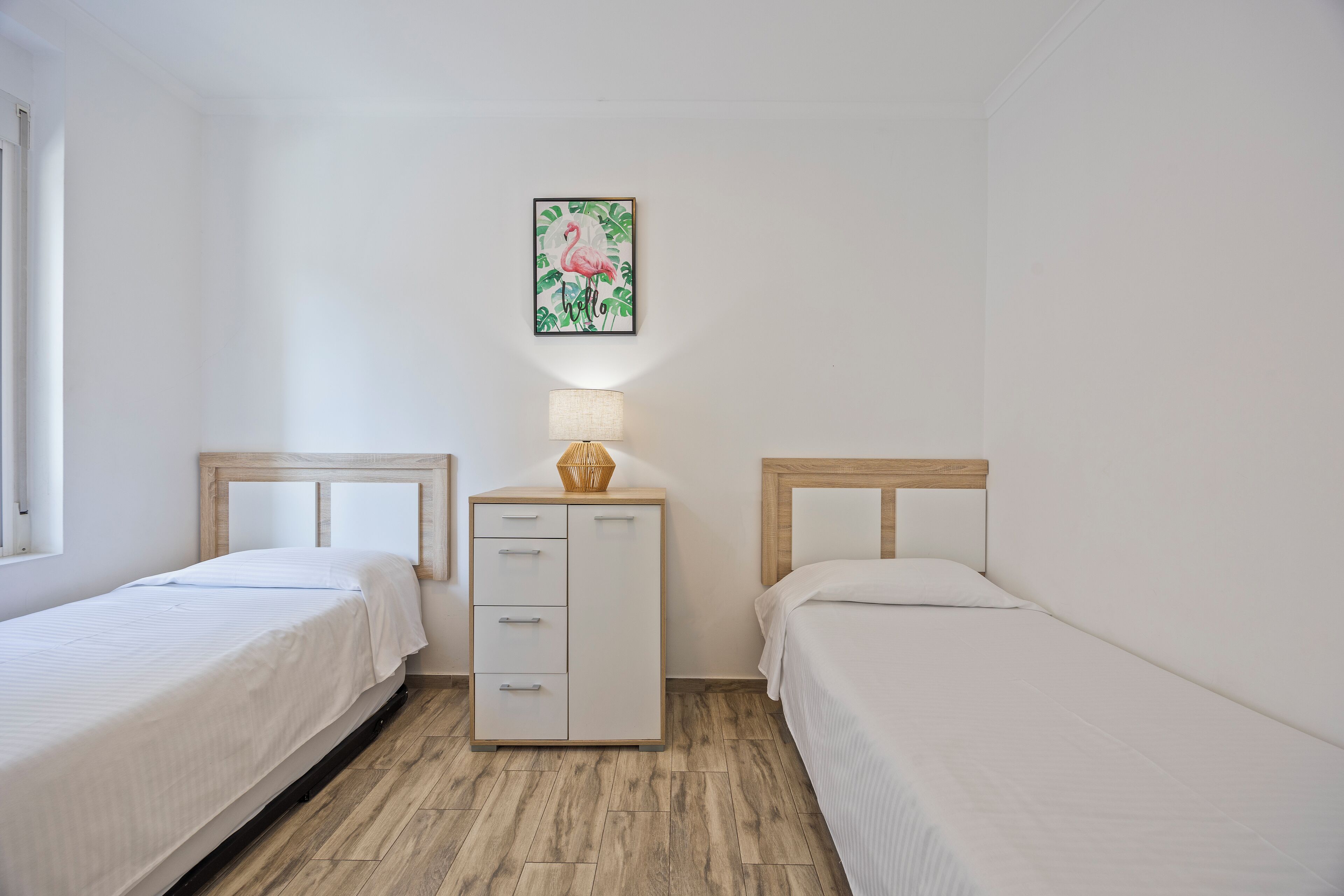 A small, clean room with two single beds, a nightstand with a lamp, and a wall-mounted picture of a flamingo.