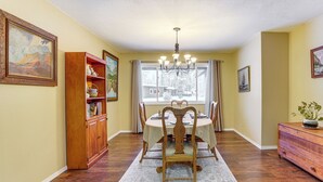 A dining area with a wooden dining table, chairs, a chandelier, and a window with curtains. - Home 20 Mi to Anchorage & Chugach State Park! (Chugiak)