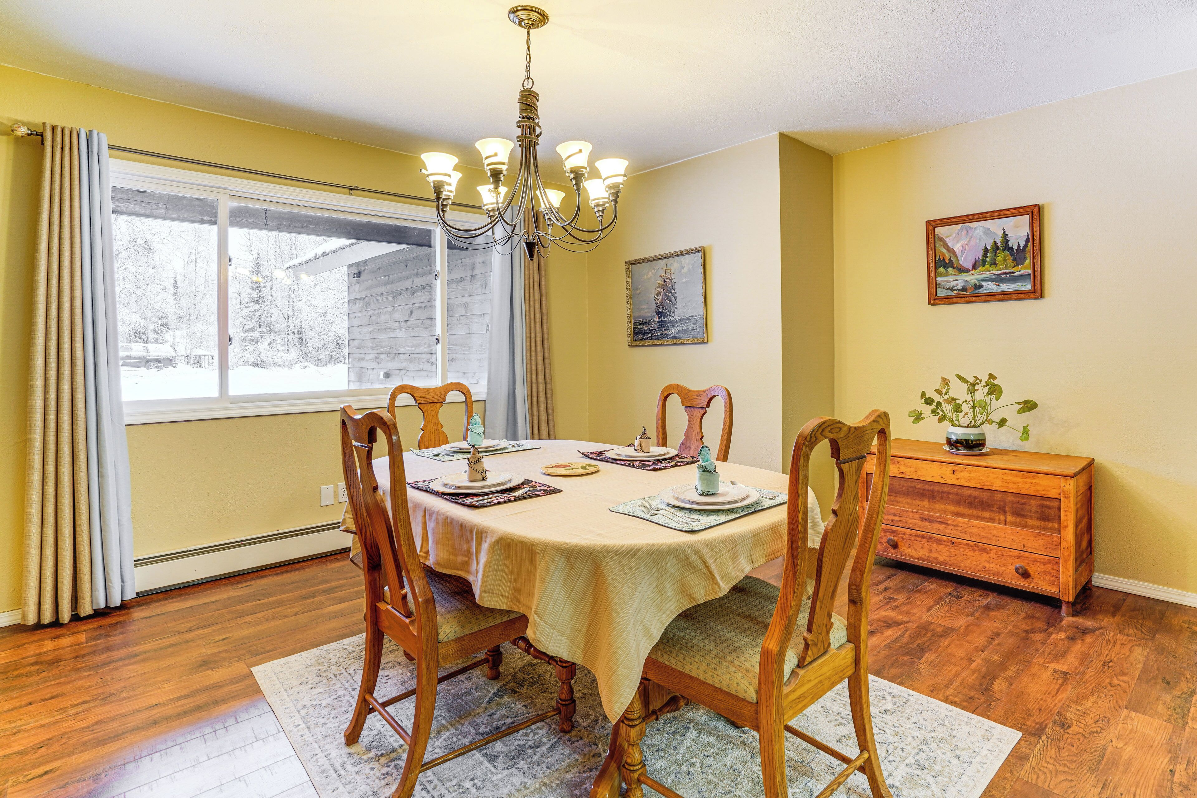 Une salle à manger avec une table en bois dressée pour quatre personnes, un lustre et une fenêtre avec des rideaux.