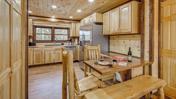 A wooden cabin interior with a kitchen area featuring wooden cabinets, a stainless steel refrigerator, a dining table with chairs, and a bench.