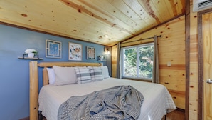 A bedroom with a wooden ceiling, a bed with a white quilt, and a window with a view of trees.