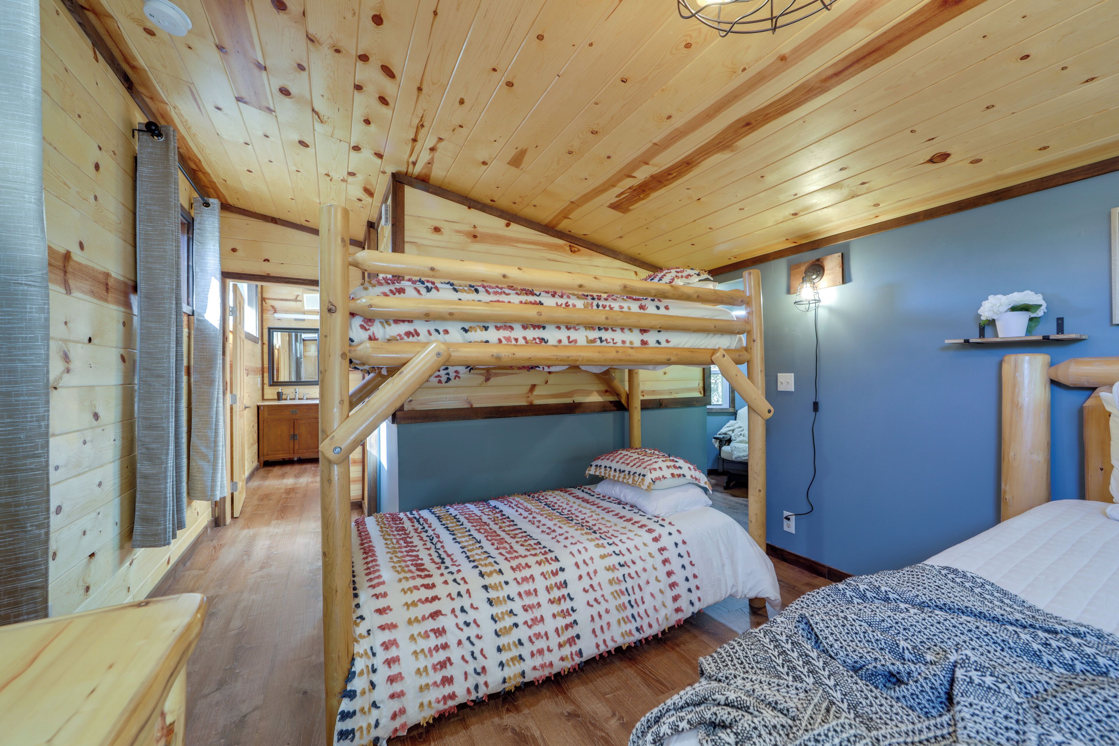 A bunk bed with a patterned bedspread, a single bed with a blue patterned bedspread, wooden walls, and a small shelf with a potted plant.