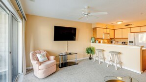 Una sala de estar con un sillón rosa, una mesa de centro de vidrio, un televisor montado en la pared y una zona de cocina con taburetes y electrodomésticos.