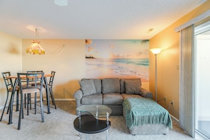 A cozy living room with a checkered sofa, a dining table with chairs, a small round table, and a painting of a beach scene. - Half Mi to Beach: Bayfront Ocean City Condo, Deck (Ocean City)