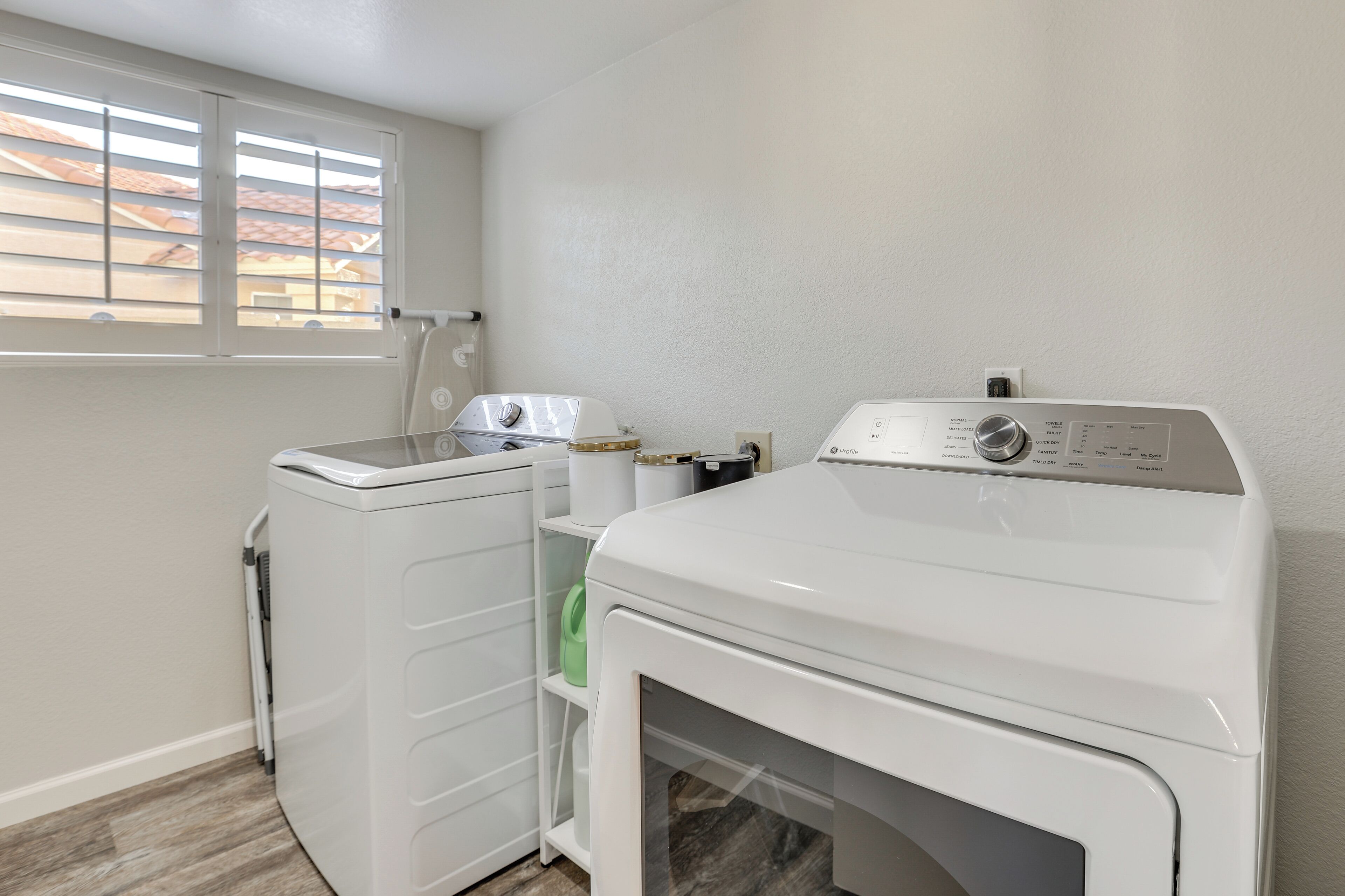 A laundry room with a front-loading washer and dryer, a window with blinds, and a shelf with laundry supplies.