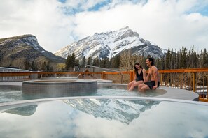 Un jacuzzi con montañas al fondo, terraza de madera y barandilla.