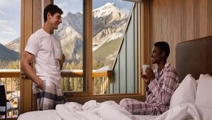 A bedroom with a large window offering a mountain view, a bed with white bedding, and a person standing by the window. - Banff Superior Queen Room w/ Incredible Vistas (Banff)