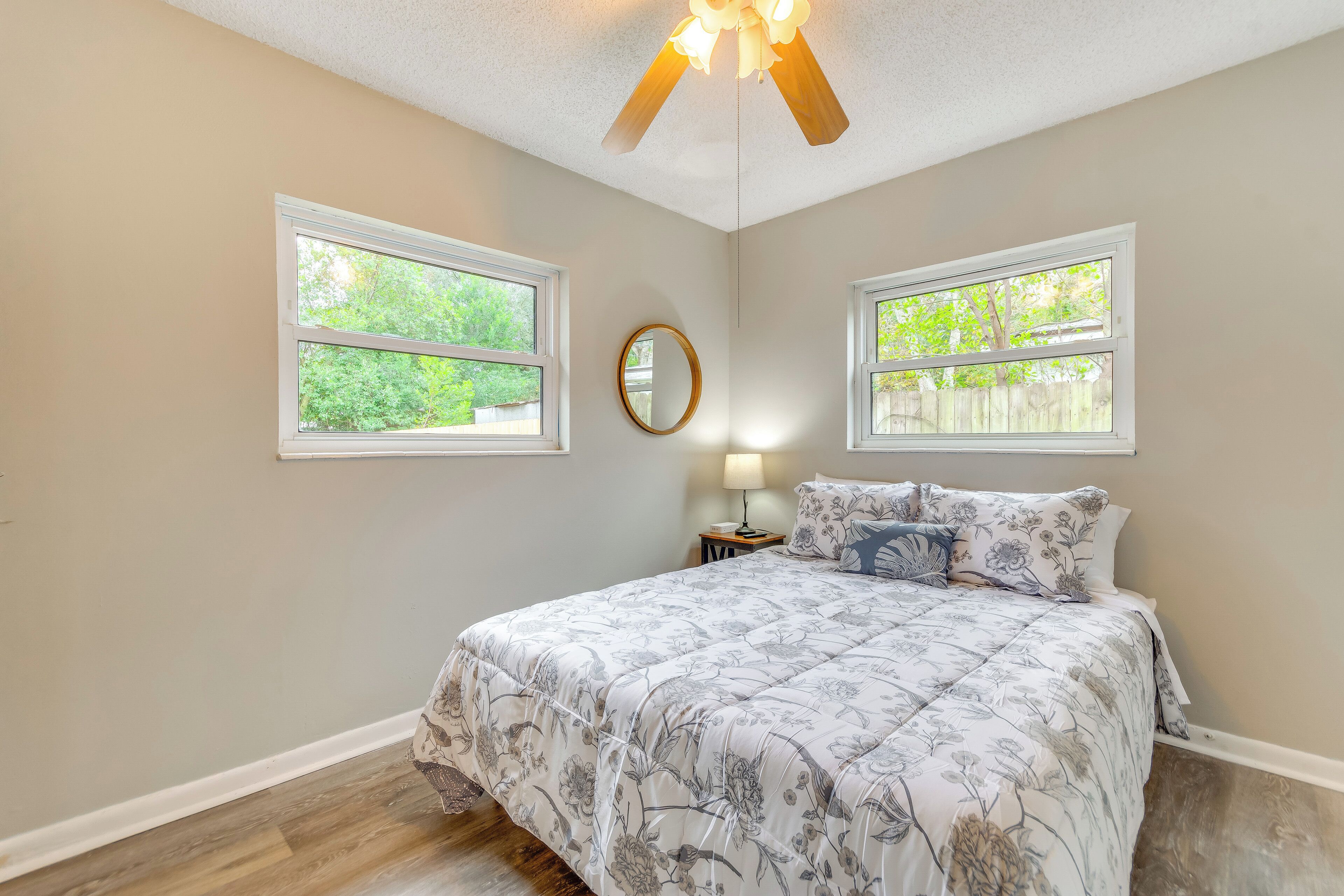 A bedroom with a large bed, two windows, a ceiling fan, and a nightstand with a lamp.