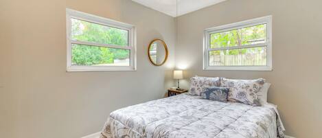 A bedroom with a large bed, two windows, a ceiling fan, and a nightstand with a lamp.