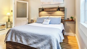 A bedroom with a bed, bedside lamps, a nightstand, a wall-mounted shelf, and a door. - East End River Retreat: Dogs Welcome! (Madison)