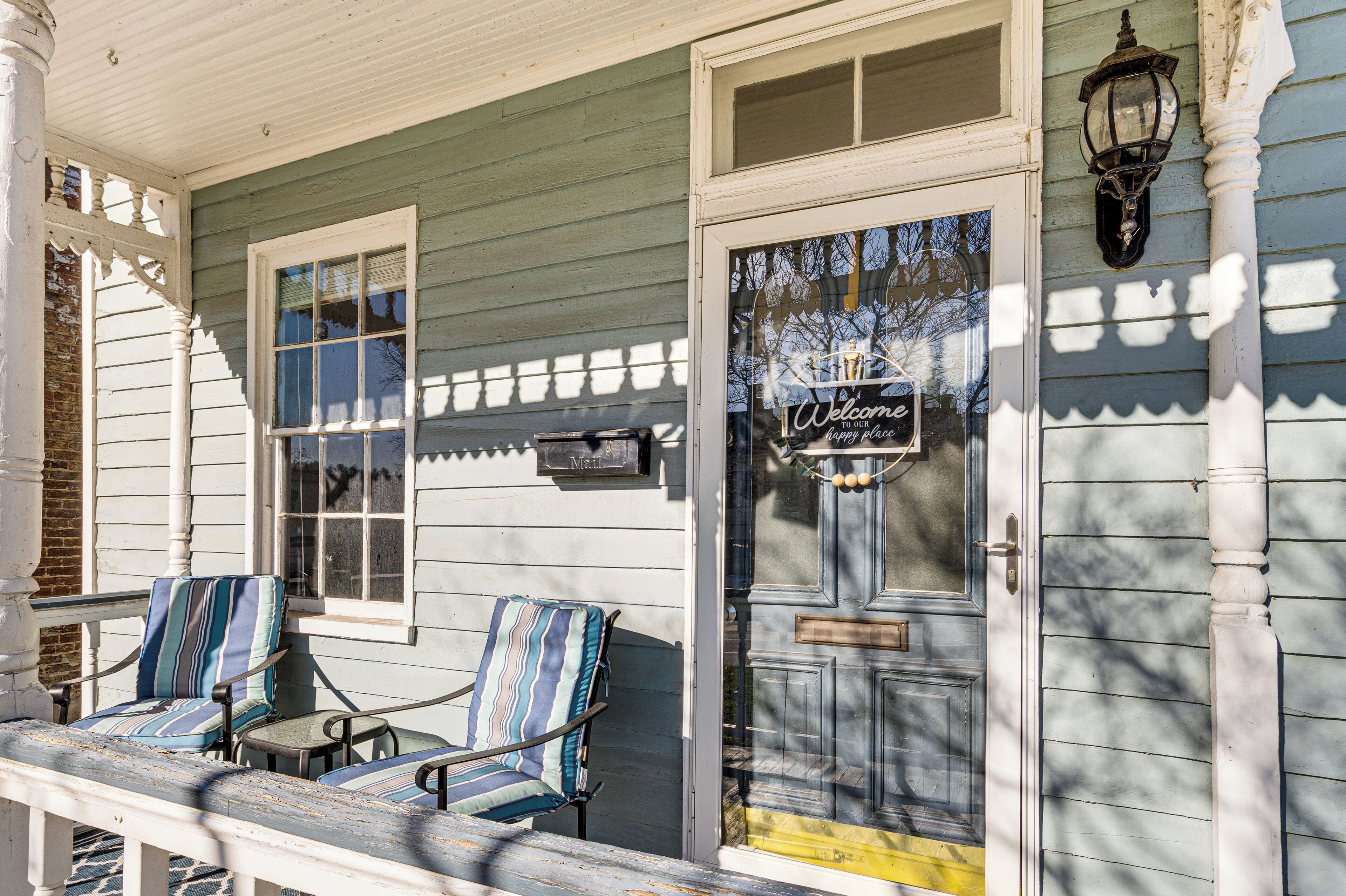 A porch with two striped chairs and a small table, a door with a 'Welcome' sign, and a wall-mounted lantern.