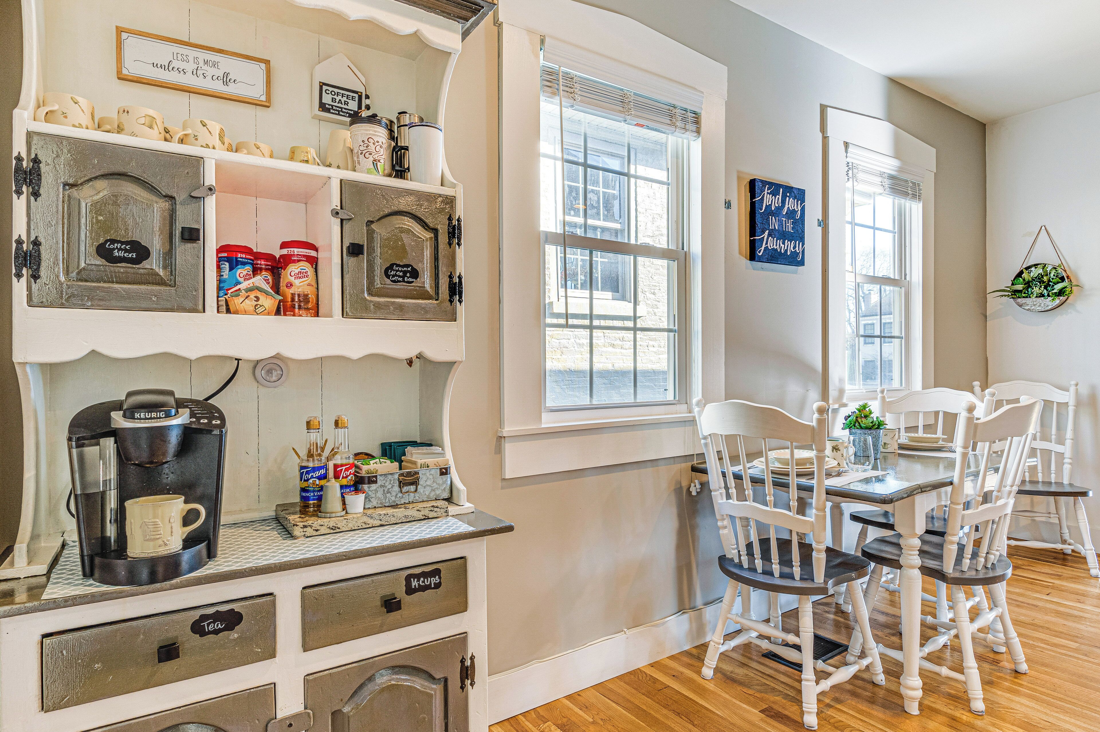 A cozy kitchen with a coffee maker, shelves with jars, and a dining area with white chairs.