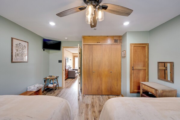 A bedroom with two beds, a wooden wardrobe, a desk, a television, and a ceiling fan. - Near Blue Mountain Lake & ATV Trails: Cabin Escape (Magazine)
