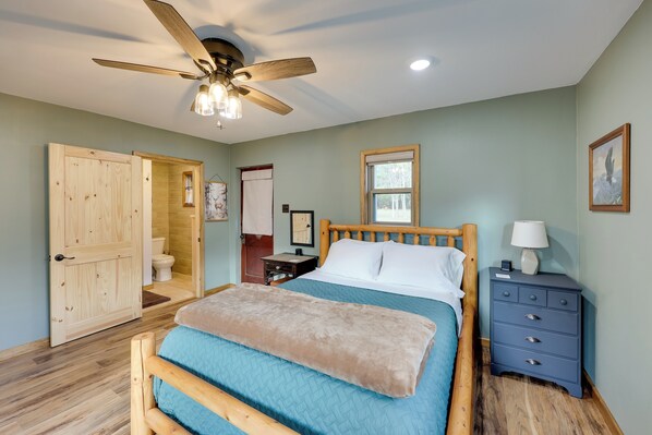 Une chambre à coucher avec un lit en bois, une table de chevet, un ventilateur de plafond et une porte en bois donnant accès à une salle de bain.