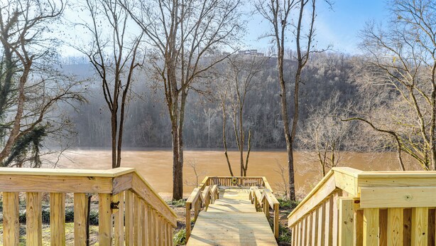 Une promenade en bois mène à une structure également en bois, dotée de rampes de protection, et est entourée d’arbres. On aperçoit une étendue d’eau en arrière-plan.