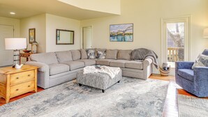 Un salon avec un canapé d’angle, un pouf et un fauteuil bleu. On y trouve une lampe, un tableau et une fenêtre donnant sur un beau paysage.