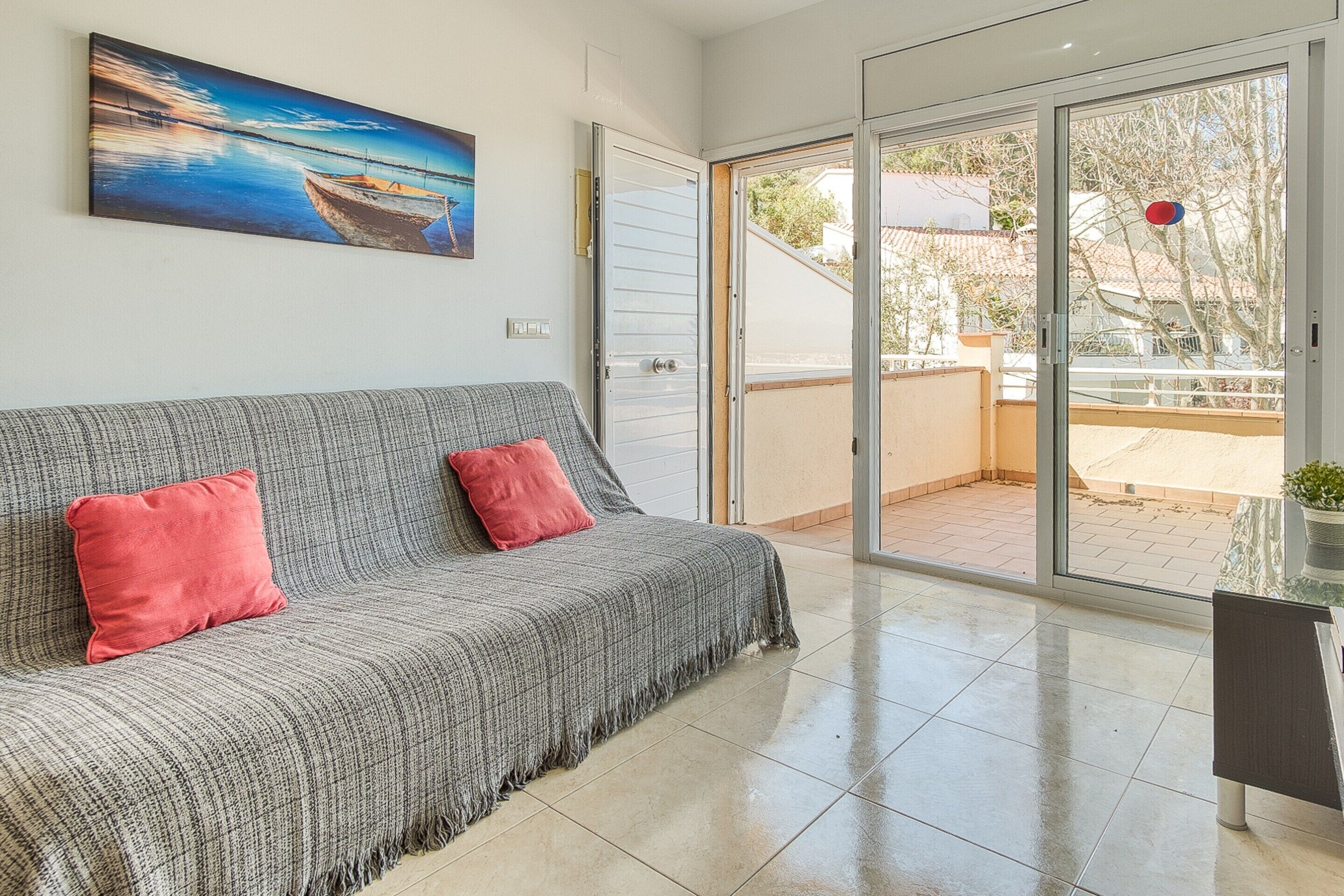 Uma sala de estar moderna com um sofá cinza, almofadas vermelhas, um quadro de um barco e uma porta de vidro deslizante para uma área externa.