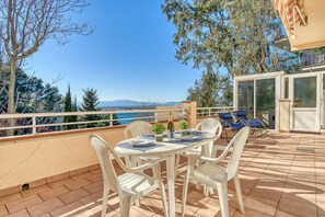 Terraço com mesa posta para dois, cadeiras de plástico branco e vista para o mar e montanhas.