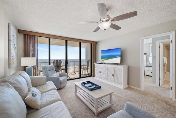A spacious living room with a large window, a ceiling fan, a flat-screen TV, and a view of the beach.