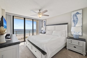 A bedroom with a bed, nightstands, a ceiling fan, and a view of the beach.