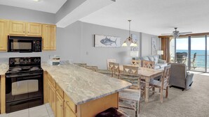 A modern kitchen with a large island, wooden cabinets, and a built-in microwave. There is a dining area with a table and chairs, and a view of the ocean through large windows. - Seaside Beach & Racquet 5817 by Liquid Life (Orange Beach)