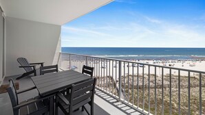Un balcon avec une table et des chaises, donnant sur une plage où l’on peut voir des gens et des parasols.