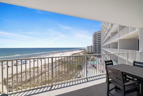 A balcony with a view of the beach and ocean, a pool area, and a dining table.