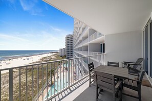 A balcony with a view of the beach and pool, a table with chairs, and a white railing.