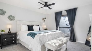 Un dormitorio con cama, mesitas de noche, un banco, ventilador de techo y ventana con cortinas.