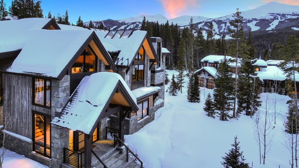 A modern lodge with multiple levels and large windows, surrounded by snow-covered trees and mountains.
