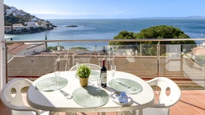 A balcony with a table set for two, overlooking a coastal view with buildings and a clear sky. - Apartment in Rosas Near Playa de Canyelles (Roses)
