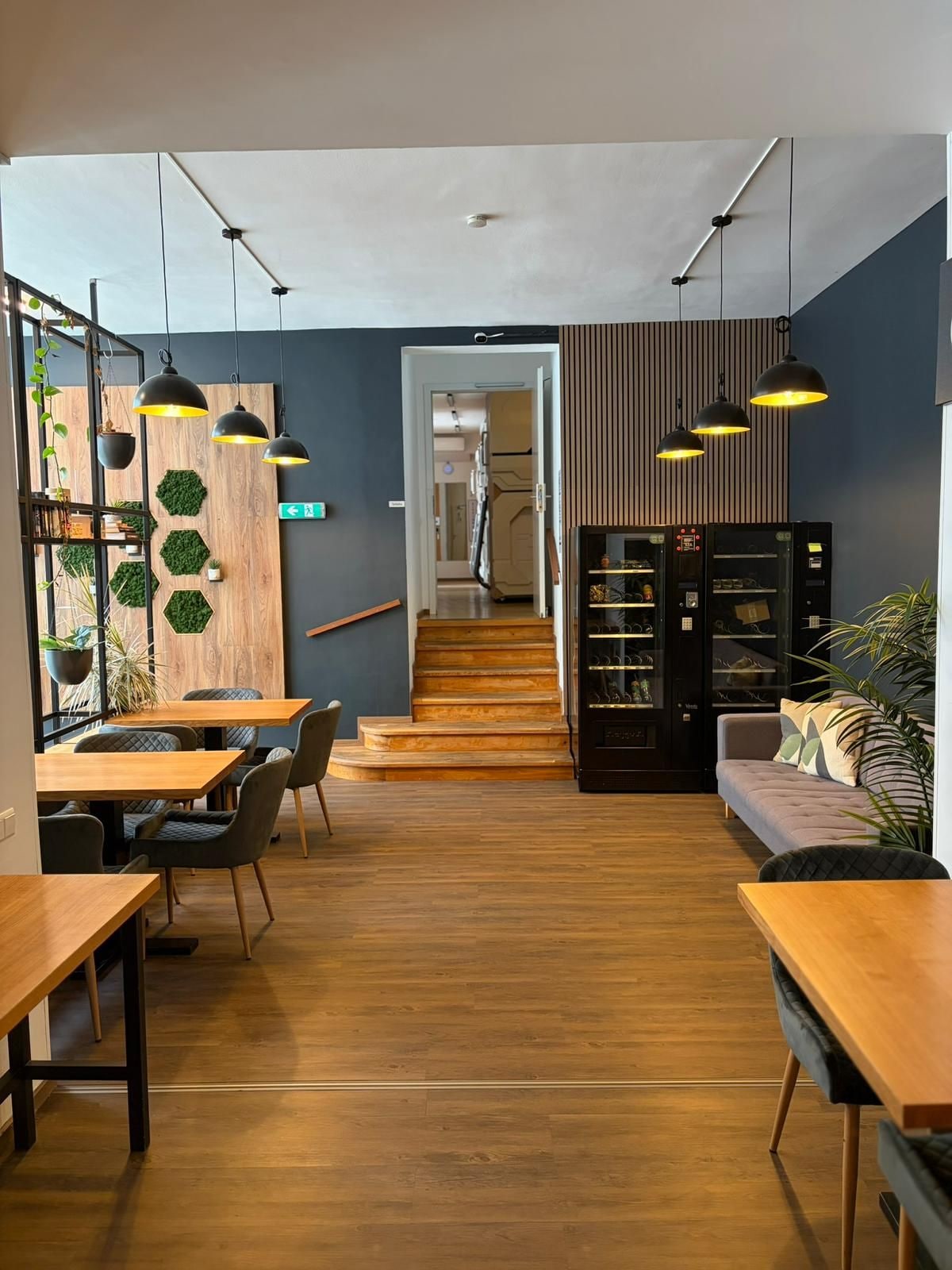 A modern interior with a wooden floor, a dining area with tables and chairs, a seating area with a sofa, and pendant lighting.