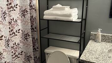 A bathroom with a toilet, a shelf with plants and towels, and a shower curtain with a leaf pattern.