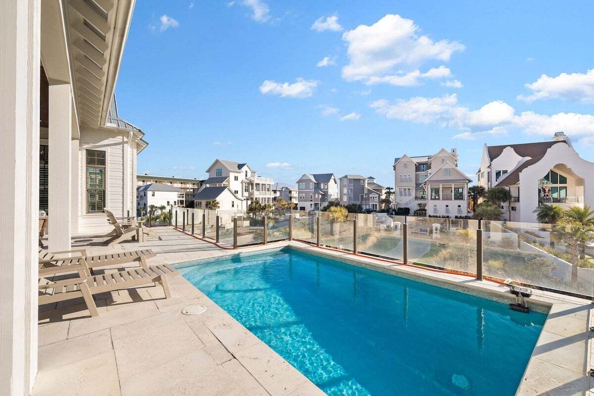 A swimming pool with a clear blue water, surrounded by a glass fence, and adjacent to a building with a white exterior.