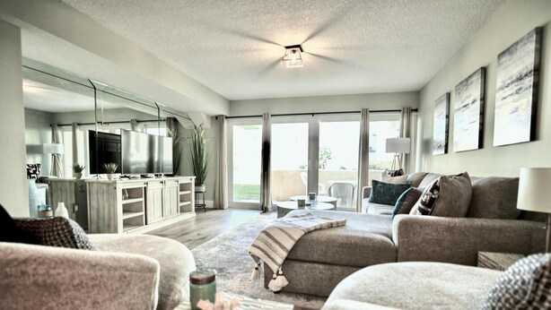 A modern living room with a sofa, a coffee table, and a TV. There is a kitchen area with a fridge and a dining table. The room has large windows and a ceiling fan.