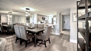 A modern kitchen with stainless steel appliances, a dining area with a large table and chairs, and a hallway leading to a bedroom.