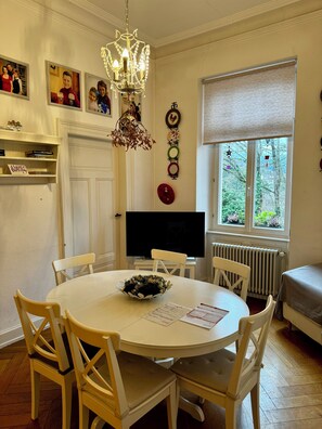 A dining area with a round table, white chairs, a chandelier, a flat-screen TV, and a window with a view of greenery. - Castle Apartment | Center Baden-Baden (Baden-Baden)