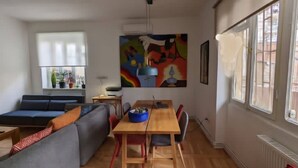 Una sala de estar moderna con un sofá, una mesa de comedor y sillas. Hay una ventana con persianas y un cuadro en la pared.