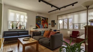 Una sala de estar con sofá, una mesa de centro y una mesa de comedor. Hay ventanas con persianas, un radiador y una lámpara en el techo.