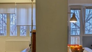 Una habitación con un ventanal grande, un radiador, una mesa con una cesta de naranjas y una silla amarilla.