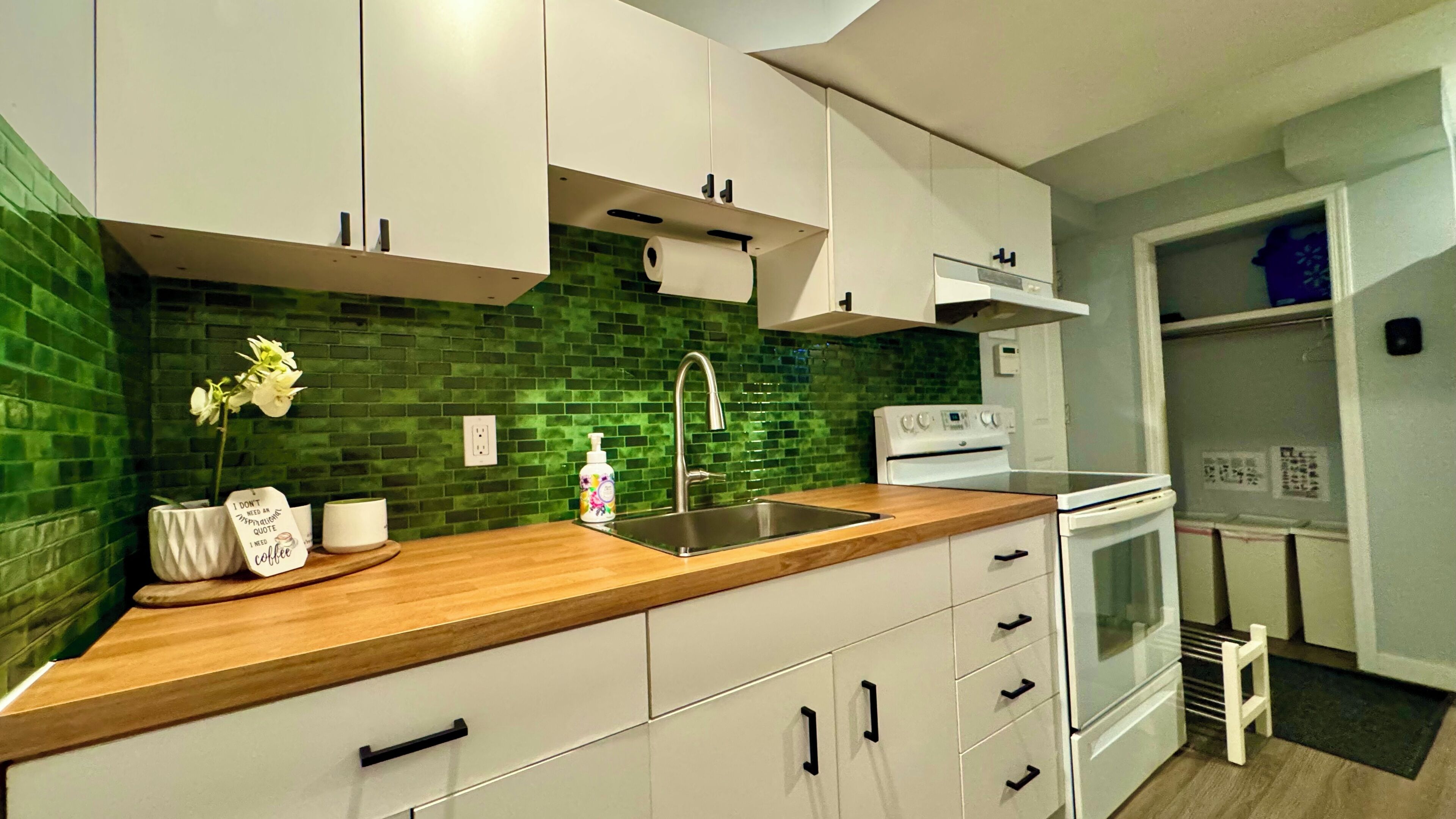 A modern kitchen with white cabinets, a green tiled backsplash, a wooden countertop, a stainless steel sink, and a white stove.