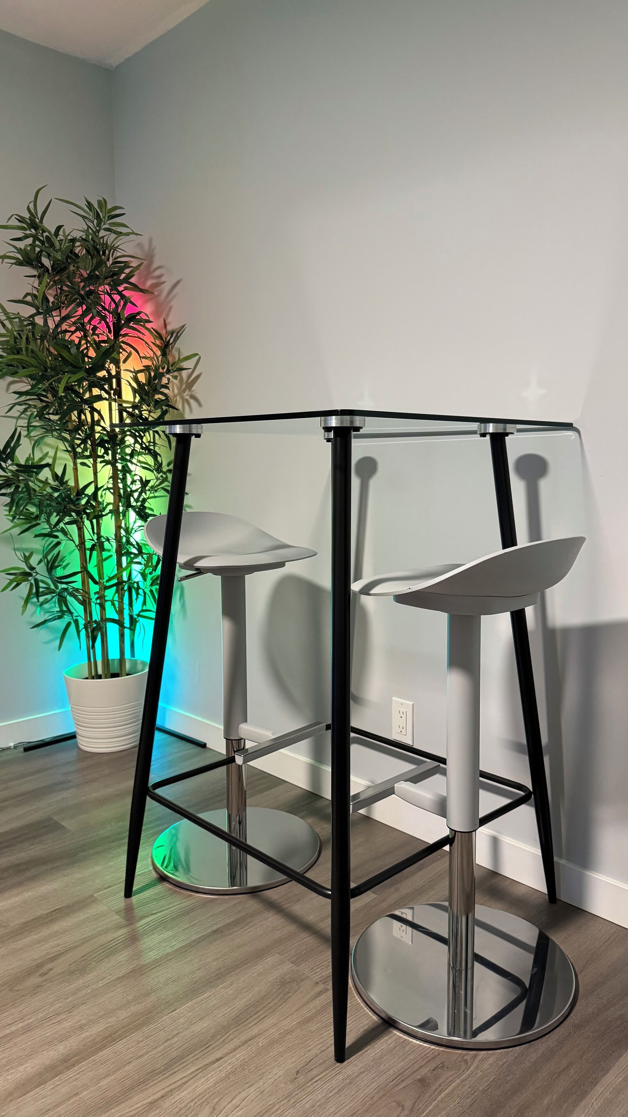 Une table moderne et minimaliste, dotée d’un cadre en métal et d’un plateau en verre, placée dans une pièce au plancher en bois et agrémentée d’une plante en pot.